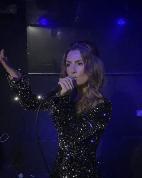 Angie North singing into a microphone in a sparkling sequinned top under blue stage light